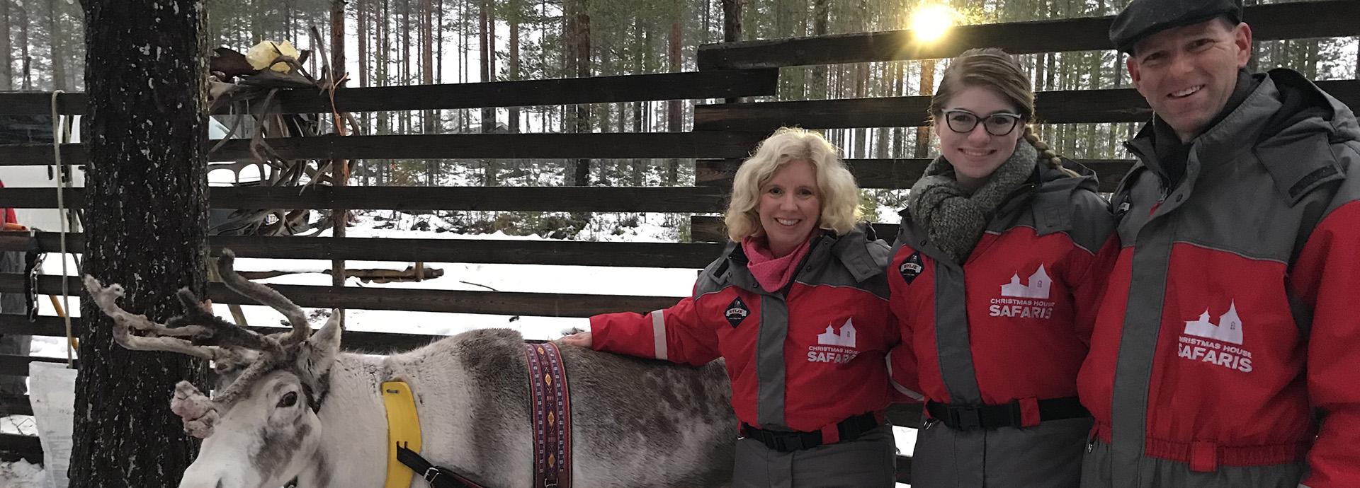 Scott Buchanan with his wife and daughter, with a reindeer on the left. The Buchanan's are wearing winter safari overalls. Behind them is a fence, through which a sun is shining. 