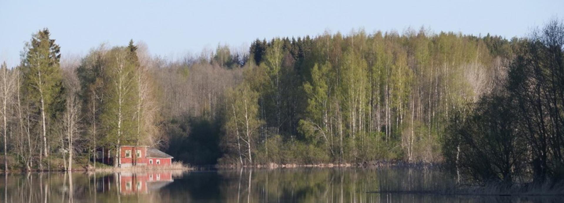 A red cottage on a shore of a still lake. There are trees behind the cottage and they are reflected in the water.