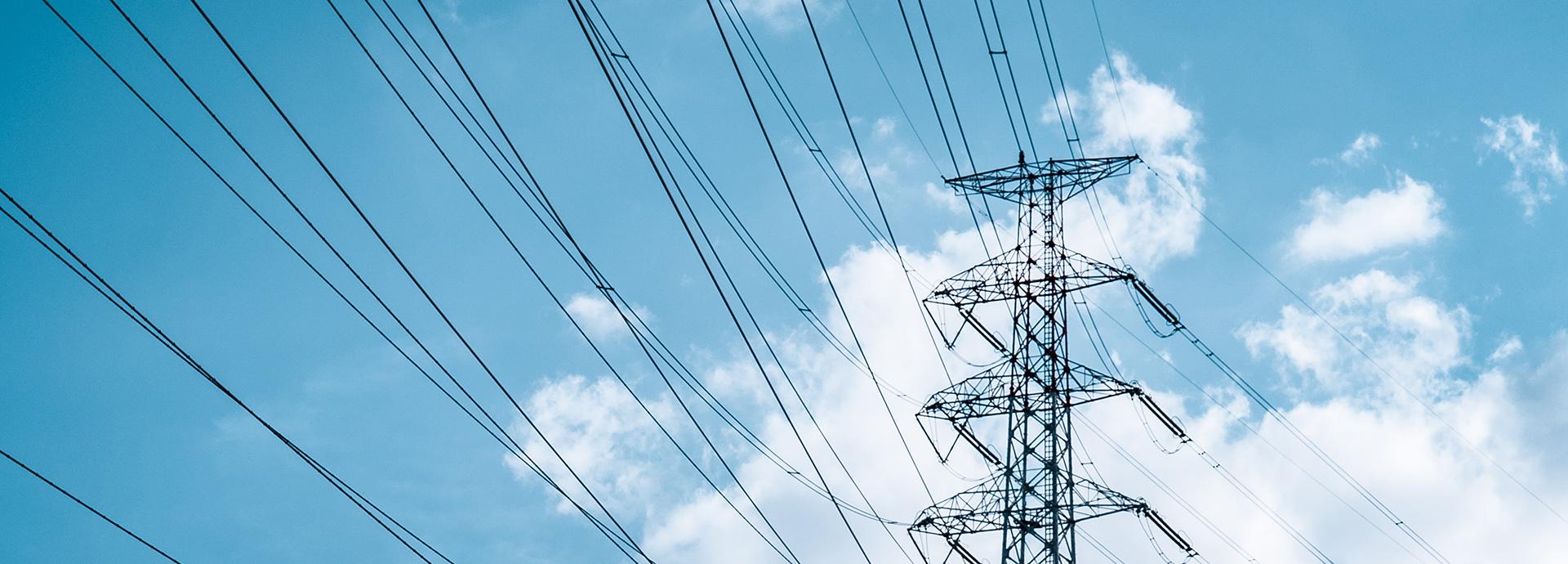 Photo of electric grid taken from down facing up, behind the tower is blue sky with white clouds