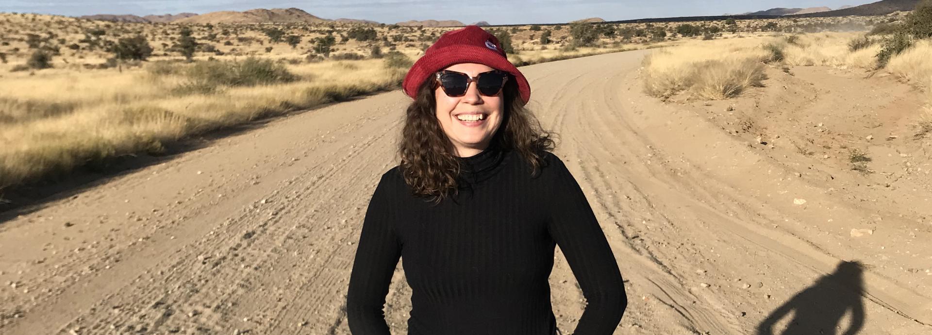 Siiri Sinnemäki standing on a deserted road on a sunny day. She's wearing a red hat and sunglasses and has a wide smile.