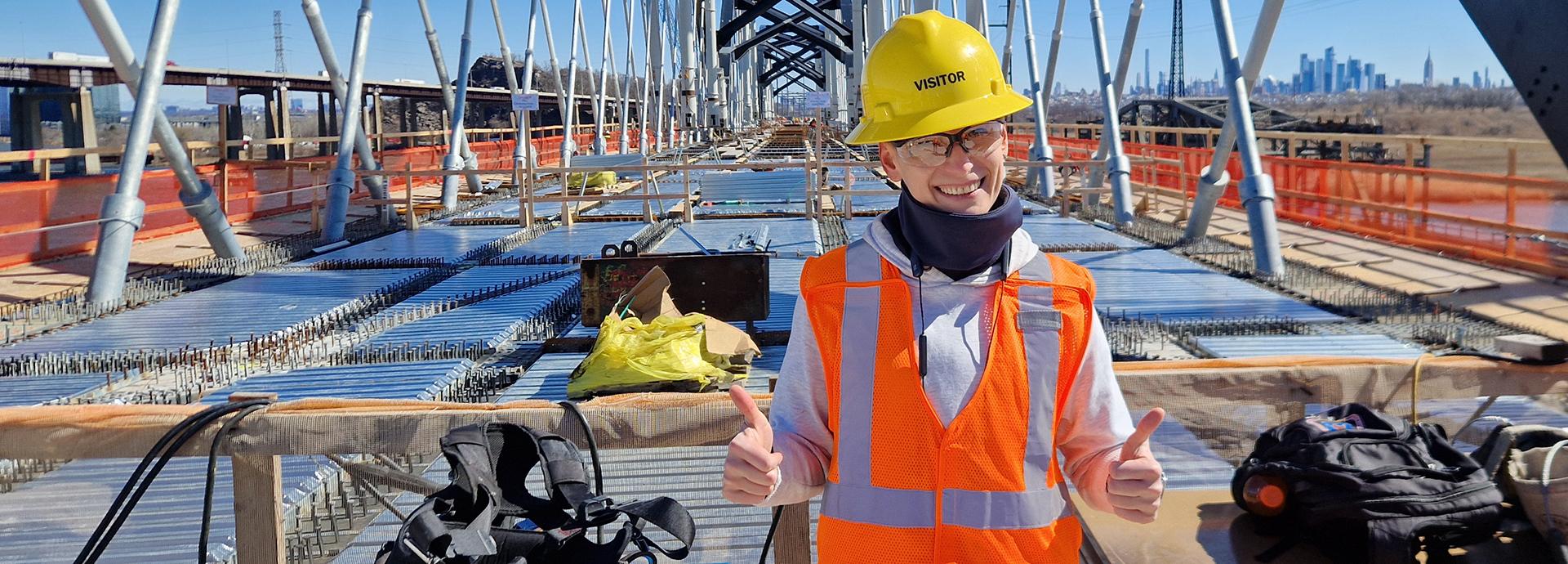 Lada Sokolova on a bridge construction site wearing a orange west, yellow hard hat and giving thumbs up to the camera