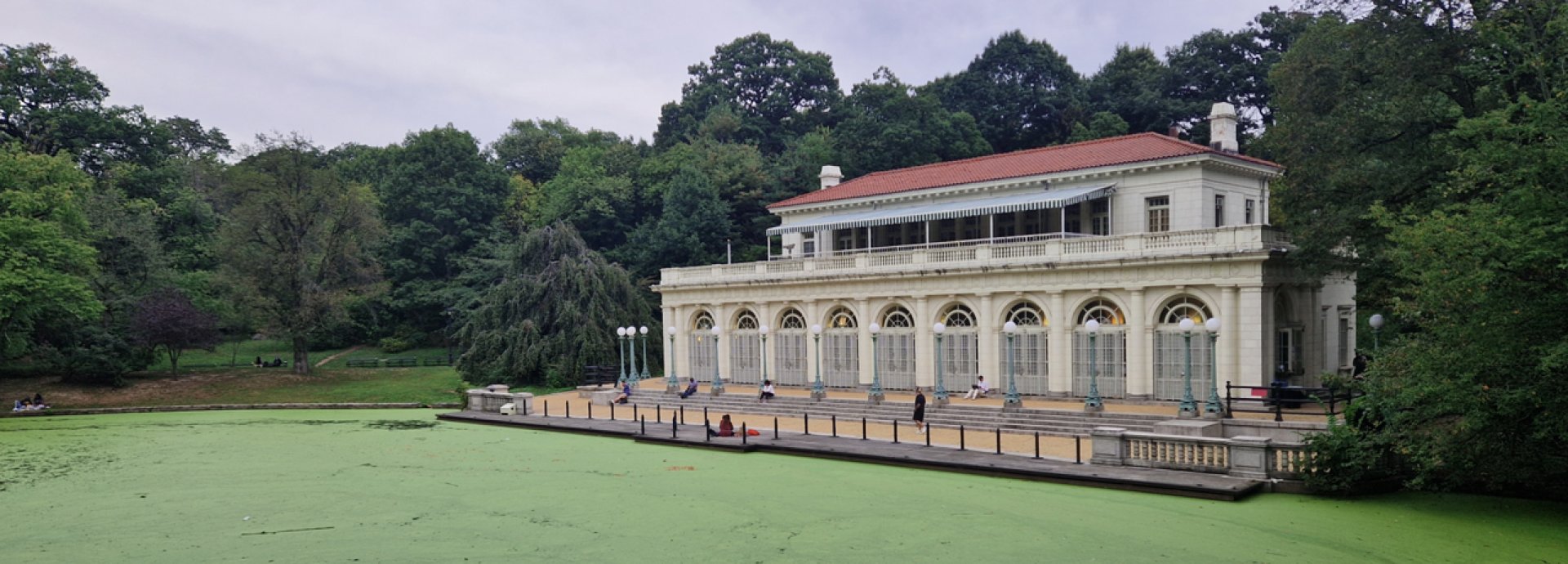 Photograph of a classical-style boathouse with arched windows and a red-tiled roof situated beside a pond covered in green algae. Surrounding dense trees and overcast sky create a serene park setting, with a few people visible on the boathouse terrace.