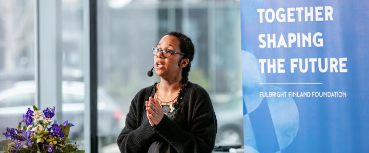 Farida Vis giving a keynote lecture in Maijansali at the Helsinki Central Library Oodi. Behind her is blue Fulbright Finland Foundation roll-up with the words Together Shaping the Future.