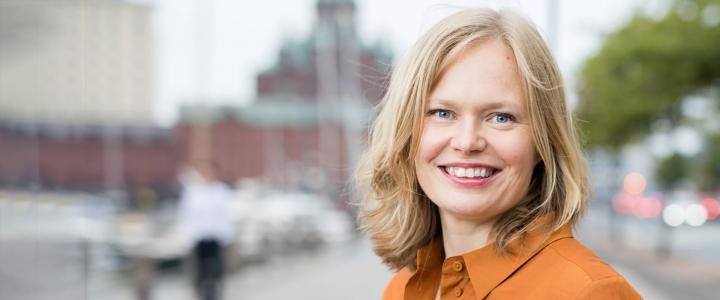 Minister of Science and Culture Hanna Kosonen photographed on a street in Helsinki, she is wearing an orange dress and is smiling at the camera.