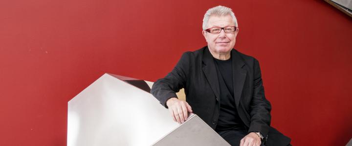 Architect Daniel Libeskind sitting on a geometrical shape with a deep red wall in the background