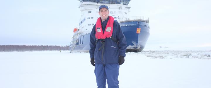 William Woityra smiling, standing on a frozen sea in front of Polaris ice breaker