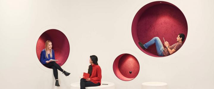 Three students hanging out in a common area where there are red circular holes in a white wall. One female student is sitting in one of the holes and talking with another student in front of her who is sitting on a low white bench. A male student is laying in one of the holes, reading a book.