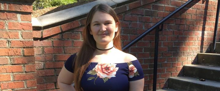 A full body photo of Johna, a young white female with red hair, standing at the landing of an outdoor staircase. She smiles, with one hand placed on her hip. She is wearing a navy blue dress with pink roses.