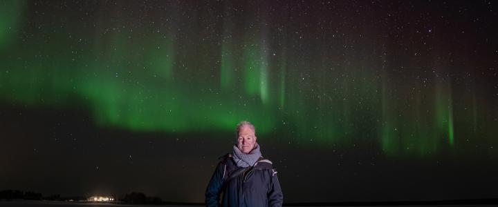 Michael West standing in the middle of the photo with green and red aurora borealis behind him.