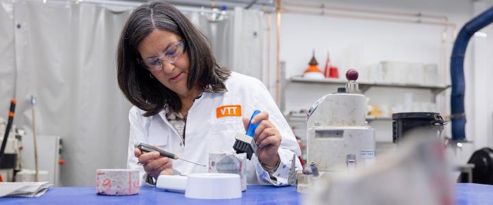 Erika Holt in a white lab coat labeled "VTT" working in a laboratory setting, using a small brush and tool on a white object. Background includes lab equipment, containers, and industrial machinery, indicating a research environment.