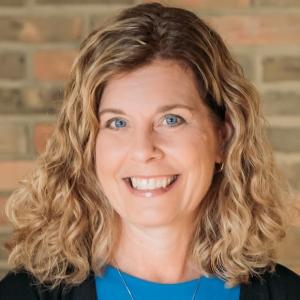 Person with blonde curly hair with a blue shirt, bricks in the background