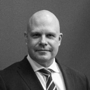 Black and white photo of a bald man wearing a black suit, white shirt and striped tie