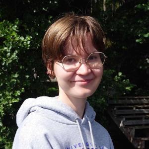 Person with short hair and wearing grey hoodie in front of greenery