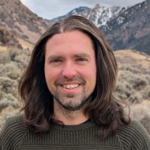 Person with shoulder length hair smiling at the camera. Mountain background