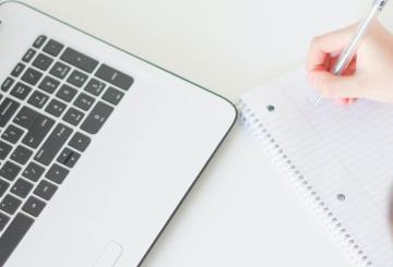 There is a corner of a laptop on the left and a person writing on a blank piece of notebook paper